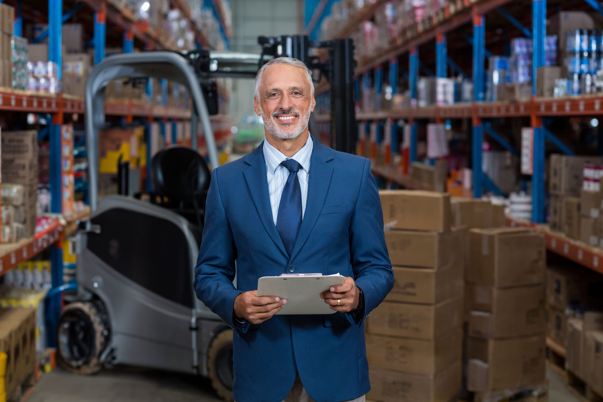 Warehouse manager standing in warehouse, holding clipboard beside forklift and shelving racks